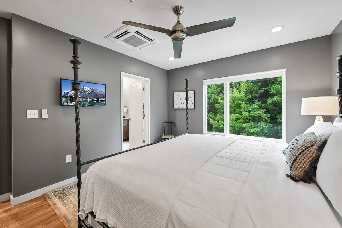 A stylish king bedroom with a prominent bed, a large window offering views of the outdoors, contemporary artwork, and a wall-mounted smart TV. The room glows with natural light and has a relaxing, cozy ambiance.