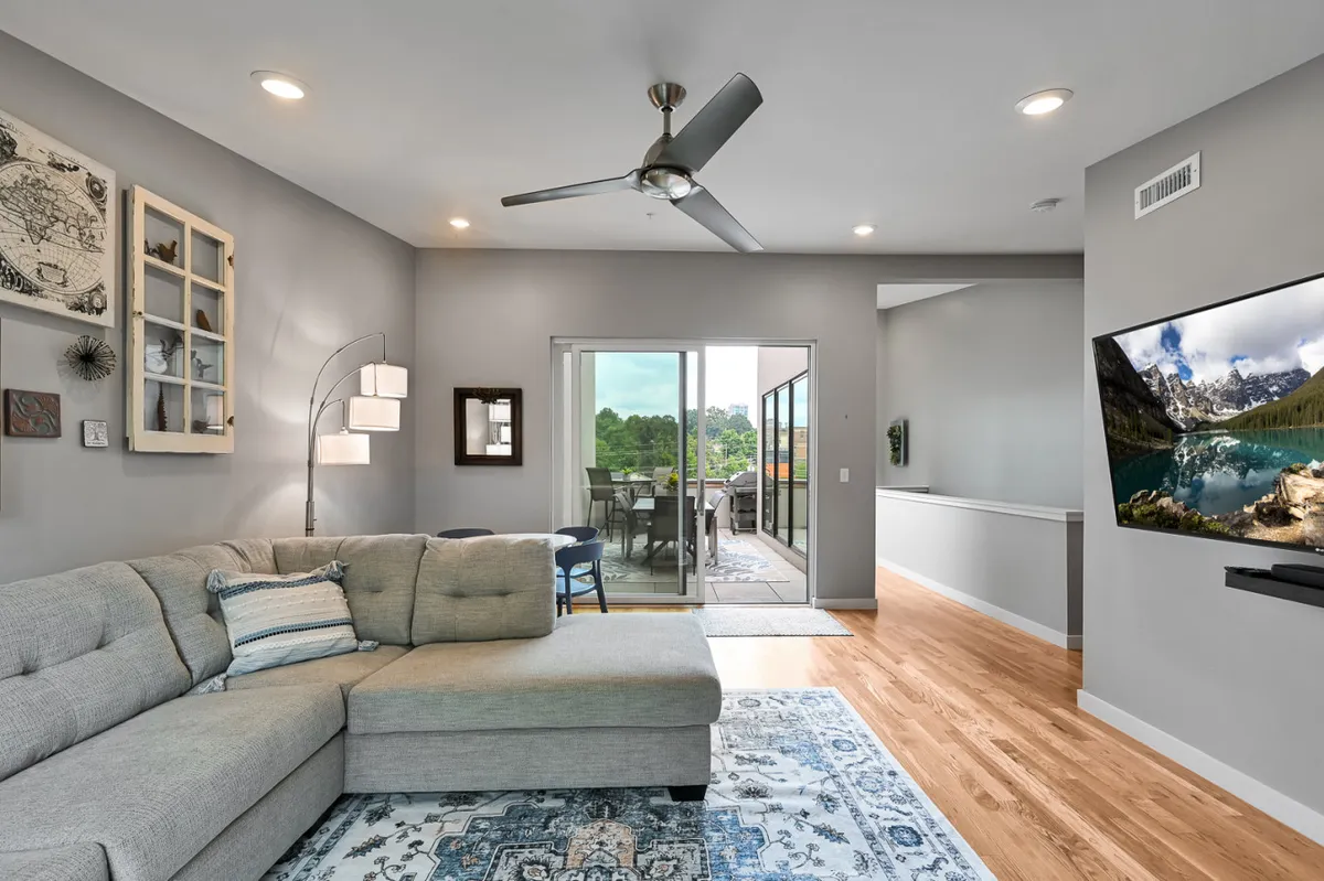 A spacious living area featuring a modern sectional sofa, vibrant wall art, a large glass door letting in sunlight, and a smart TV setup. The space feels open, bright, and perfect for lounging, movie nights, or casual gatherings.