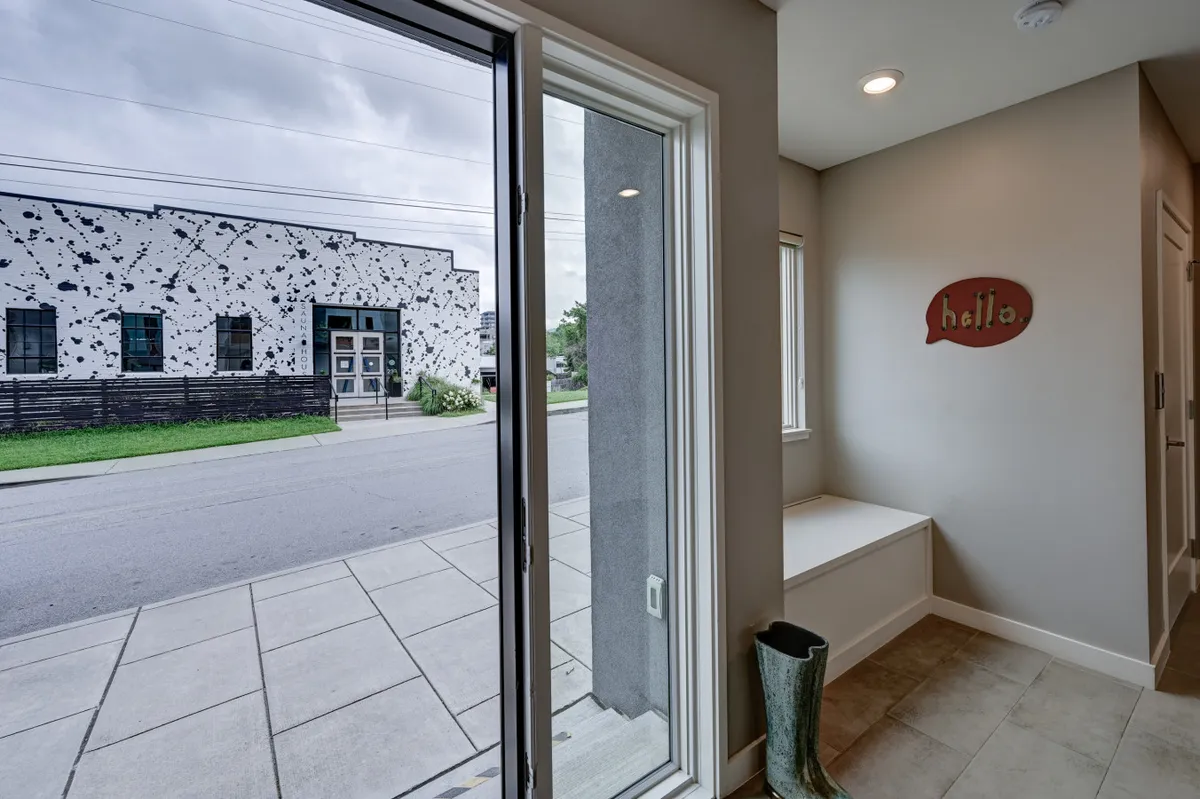 A bright, inviting entryway with a large window, a stylish bench, a decorative 'hello' sign, and a pop of color from green rain boots. The scene is bathed in natural light, conveying a welcoming atmosphere and city living charm.