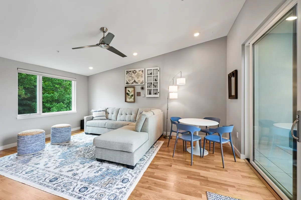 A bright, multifunctional interior area with a cozy sofa, area rug, dining table, and large windows allowing plenty of natural light. The arrangement encourages both relaxation and social activities, suitable for reading, socializing, or creative pursuits.