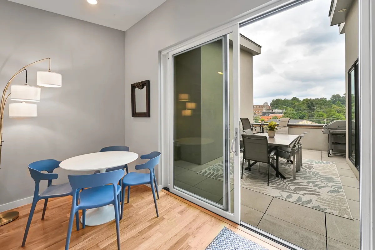 A bright dining area with blue chairs around a table, adjacent to a sliding glass door that opens to an outdoor terrace featuring comfortable seating and a view of the city beyond. The space is filled with natural light and offers an inviting transition from indoors to outdoors.